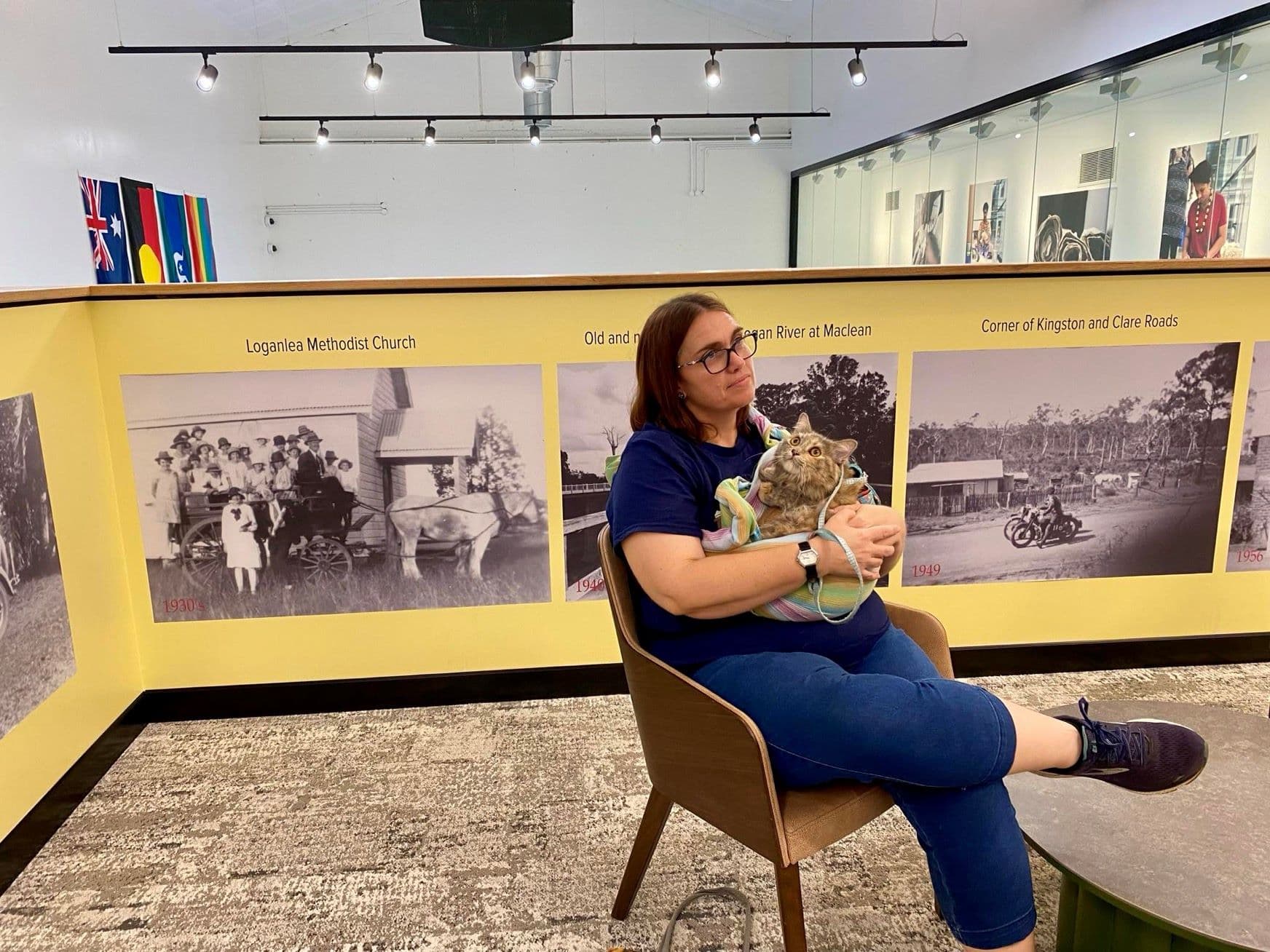 A woman sits in a gallery, holding a small dog before historic photo displays. A woman sits in a gallery, holding a small dog before historic photo displays.